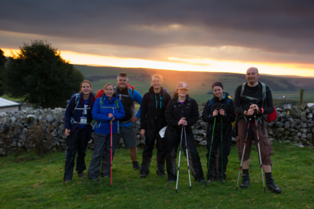 A smaller group of climbers smiling at the camera at the top of Snowdon with the sun rising behind them.