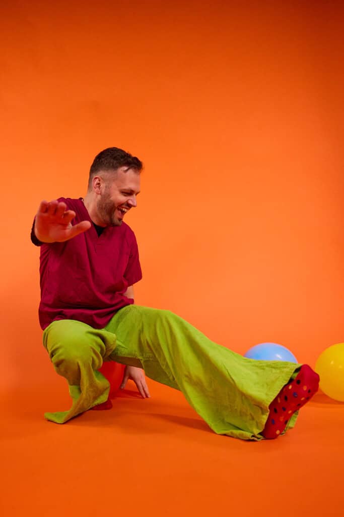 Young man dressed in work hospital scrubs getting involved in a fundraising event to raise money for sick kids. The man is wearing red scrubs from the children's hospital, green wrong trousers against an orange background. The photo is showing you can get involved in Wallace & Gromit's Wrong Trousers Day from your work, office, hospital or company.