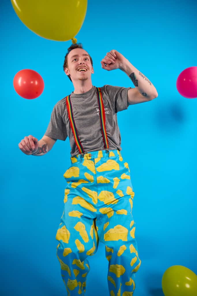 Young man is dressed in office clothing and wearing wrong trousers that include cheese. Man is wearing a grey work shirt, non-uniform trousers against a blue background. The photo is showing people can get involved in Wallace & Gromit's Wrong Trousers Day at work.