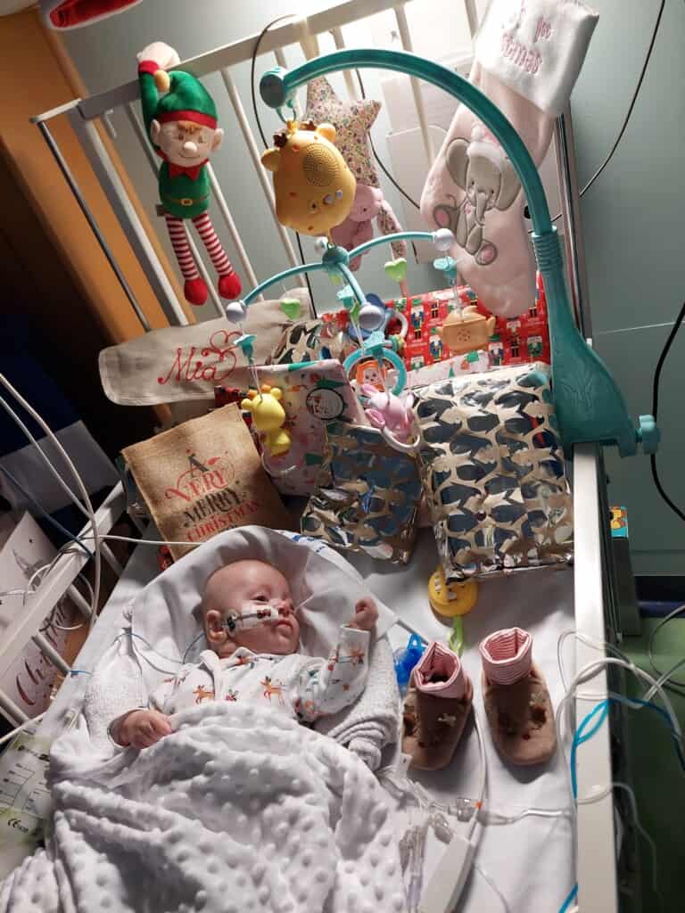 Donate this Christmas to help children like Mia. Mia is in a cot with presents in this photo.