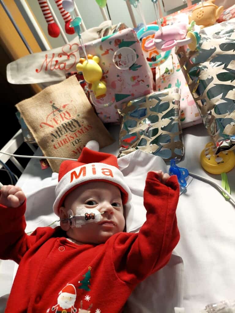 Donate this Christmas to help children like Mia. Mia is in a cot with presents in this photo.