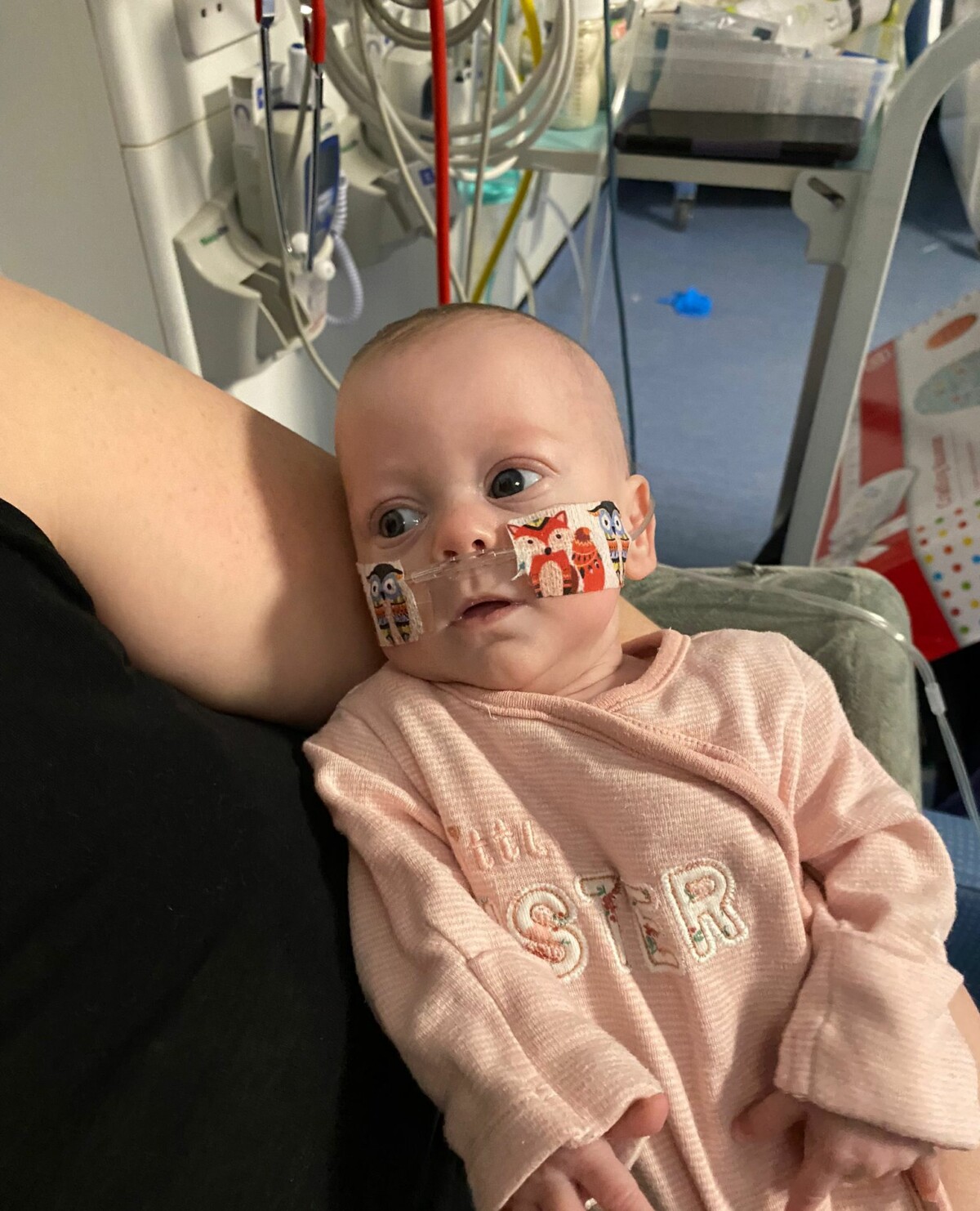 Jade sits in her mum's lap, looking away from the camera, wearing a pink onesie and has a feeding tube in.