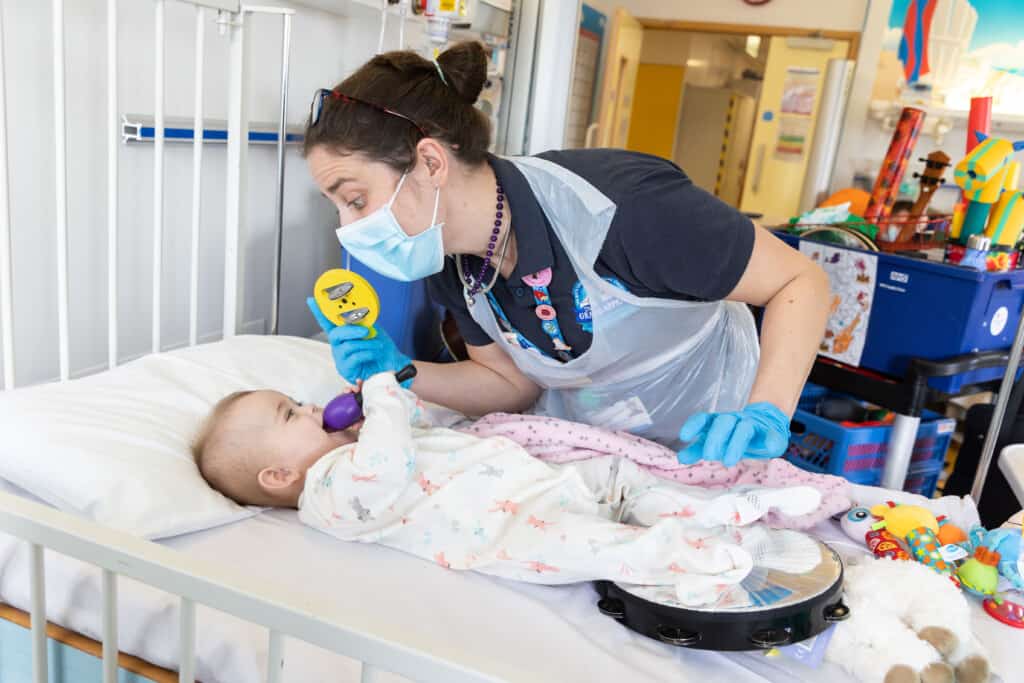 A smiling baby girl in a hospital bed playing with one of The Grand Appeal's Music Therapists.