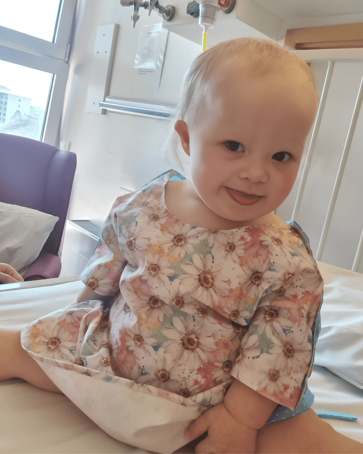 A toddler, wearing a hospital gown with daisies on it, grins at the camera.