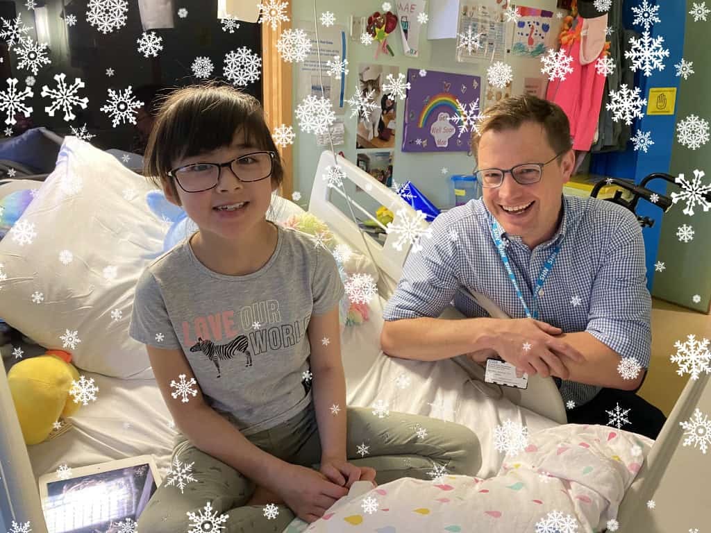 Mae sits on the bed with her doctor and smiles at the camera. There are snowflakes on the image.