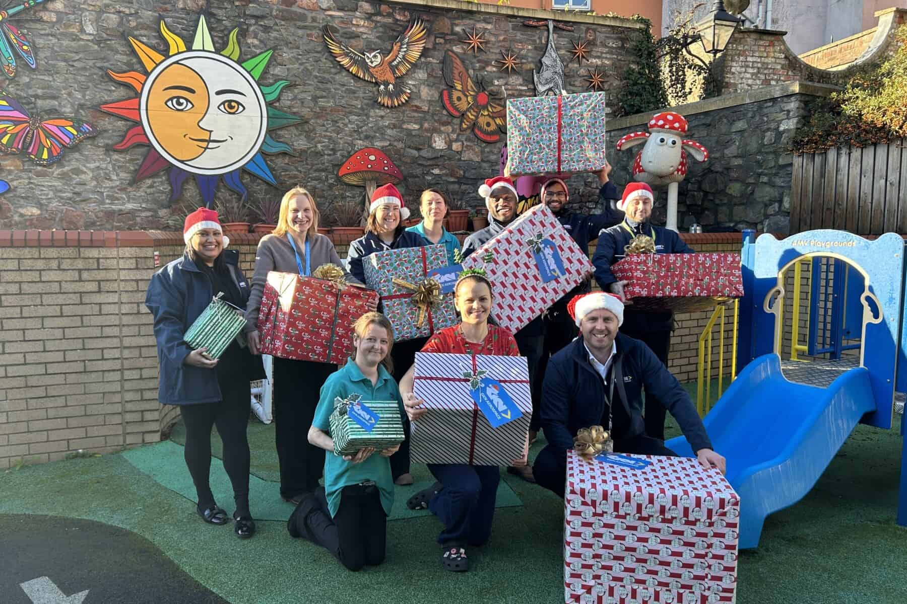 GKN Aerospace and the Bristol Children's Hospital Play Team stand in a sunny garden. They are wearing Santa hats and holding Christmas presents.